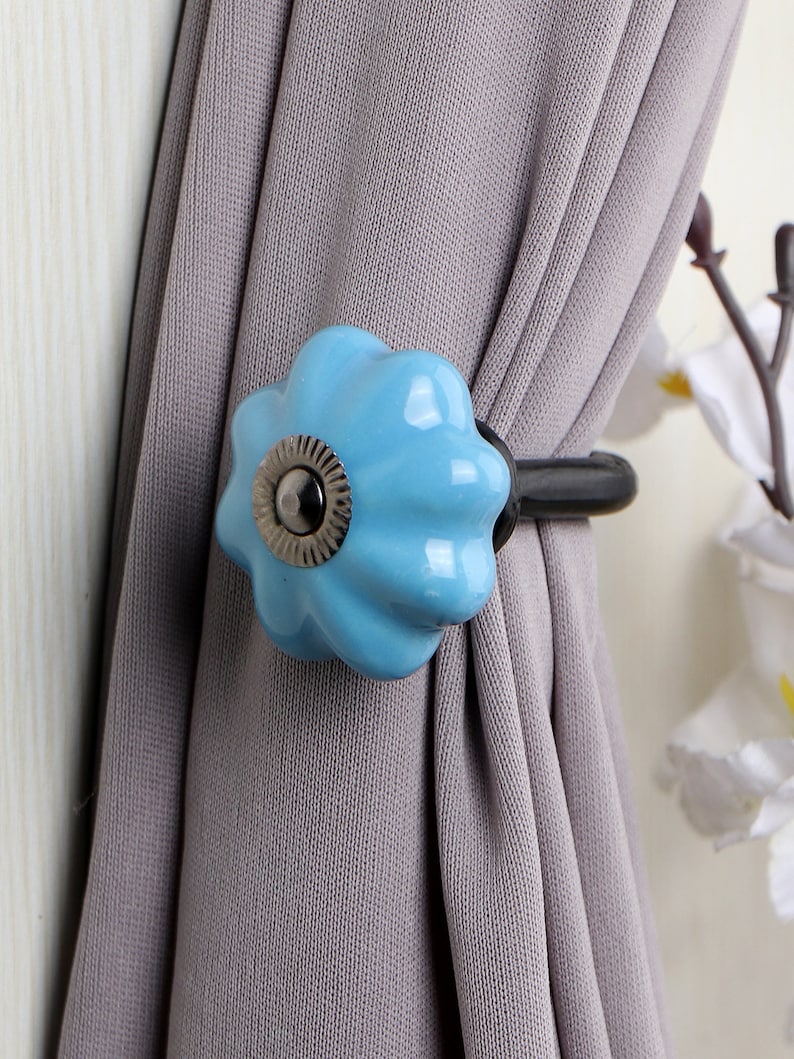 May include: A blue ceramic curtain tieback with a silver knob and a black metal hook. The tieback is attached to a gray curtain.