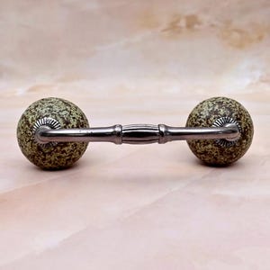 May include: A cabinet handle featuring two spherical knobs with a speckled green and brown pattern, connected by a horizontal metallic bar. The bar has a central ribbed section and silver-toned hardware where it attaches to the round knobs.