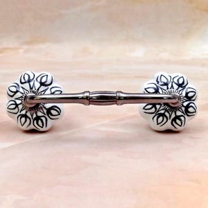 May include: A drawer pull featuring two white ceramic knobs with a black floral pattern and a scalloped edge. The knobs are connected by a smooth, metallic, silver-toned bar that has a central decorative band and fluted circular bases.