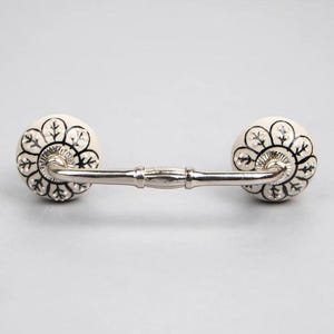 May include: White ceramic drawer pull with a black floral design. The pull has a silver metal bar connecting the two knobs.