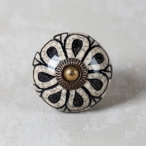 以下が含まれることがあります： 黒と白の花柄が施された白いセラミック製のノブで、中央には金色の金属製の装飾があります。