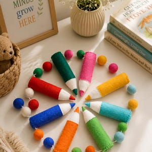 May include: A circular garland featuring seven felt pencil-shaped ornaments in red, blue, orange, green, teal, yellow, and pink, each with a white tip. The pencil ornaments are connected by a string interspersed with small, multicolored felt balls.