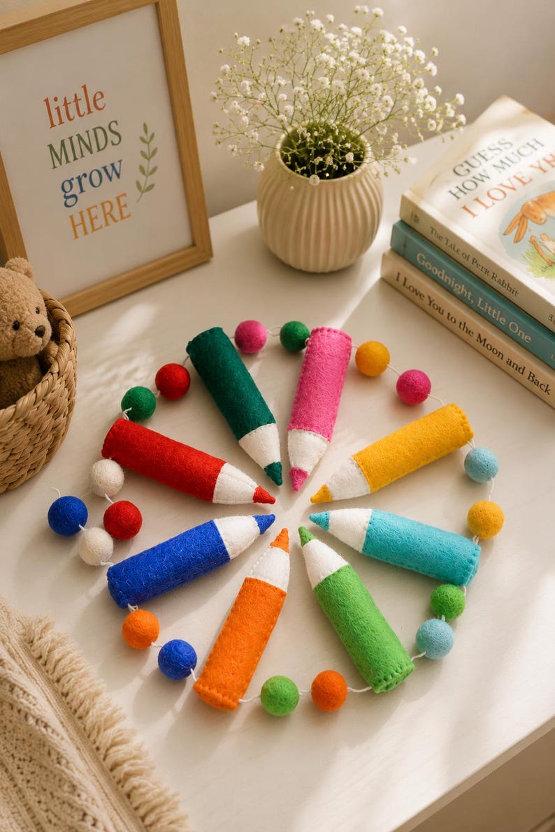 May include: A circular garland featuring seven felt pencil-shaped ornaments in red, blue, orange, green, teal, yellow, and pink, each with a white tip. The pencil ornaments are connected by a string interspersed with small, multicolored felt balls.