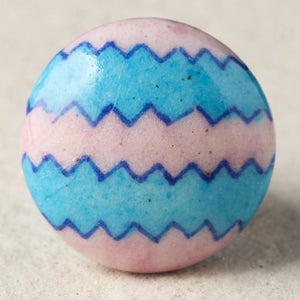 May include: A round ceramic knob with a pink base and two blue zig-zag stripes. The knob has a glossy finish.