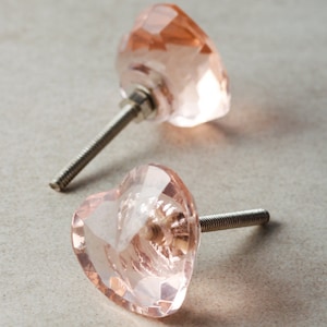 May include: Two faceted, heart-shaped cabinet knobs in a soft pink hue. Each knob features a silver-colored threaded post for mounting. The knobs are made of a clear, possibly crystal, material.