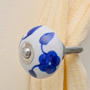 Puede incluir: Recogedor de cortina floral blanco y azul. El recogedor de cerámica presenta una base redonda blanca con diseños florales azules y un anillo de metal plateado. Sujeta una cortina color crema.