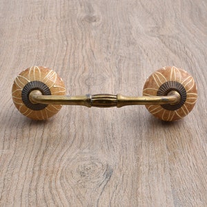 Puede incluir: Un tirador decorativo para armario con dos pomos redondos de cerámica color beige claro con un diseño floral blanco. Los pomos están conectados por una barra metálica color latón con una sección central con aspecto de madera. La ferretería está sobre un fondo claro de vetas de madera.