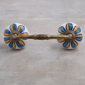 以下が含まれることがあります： 青と黄色の花柄のセラミックノブが2つ付いた装飾的な引き出しの取っ手。ノブは真鍮の棒でつながっています。