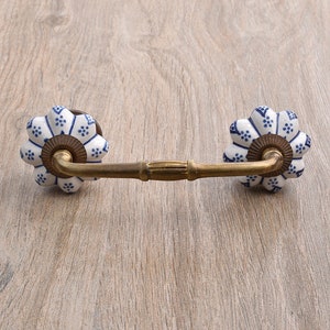 Puede incluir: Un tirador de cajón de cerámica blanca con un patrón floral azul y una barra de metal dorado que conecta los dos pomos.