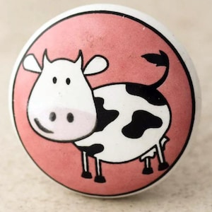 May include: A white ceramic knob with a black and white cow design on a pink background. The cow is facing the right side of the knob.