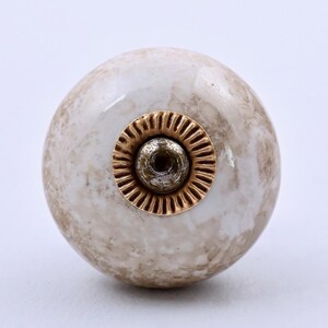 May include: A round cabinet knob with a glossy, marbled finish in shades of beige, cream, and white. The center features a circular, metallic gold-toned accent with a ridged, sunburst-like pattern surrounding a central screw hole for mounting.