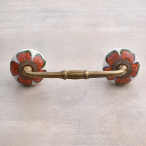 May include: A brass drawer pull with two ceramic knobs. The knobs are white with a red and green floral design.