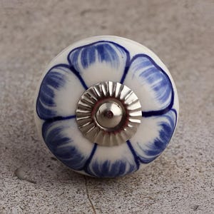 May include: A white ceramic cabinet knob with a blue floral design. The knob has a silver-colored metal base and a small, rounded center. The floral pattern is painted in shades of blue, creating a decorative accent for furniture.