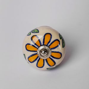 May include: A white ceramic knob with a yellow flower and green leaves painted on it. The knob has a silver metal screw in the center.