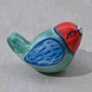 Peut inclure: Une petite figurine d'oiseau en céramique avec un corps vert, des ailes bleues et une tête rouge. L'oiseau a un œil blanc et une pupille noire.