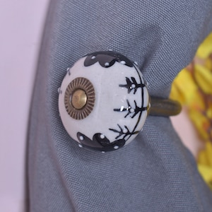Könnte beinhalten: Ein dekorativer Gardinenhalter mit einem weißen Keramikknauf mit schwarzem floralem Design. Der Knauf hat eine Messingmitte und ist an einem bronzefarbenen Metallring befestigt. Er hält einen grauen Vorhang.