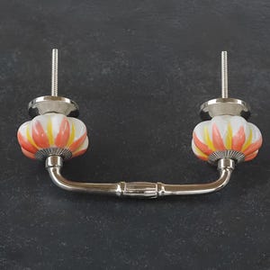 Könnte beinhalten: Ein silberner Schubladenzug mit zwei Keramik-Knöpfen in Blumenform. Die Knöpfe sind weiß mit orangefarbenen und gelben Akzenten.