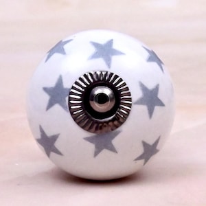 Op de afbeelding: Een ronde witte keramische meubelknop met een herhalend patroon van grijze sterren. Het midden van de knop is voorzien van een cirkelvormig zilverkleurig metalen beslag met een geribbelde rand en een centraal schroefgat voor montage.