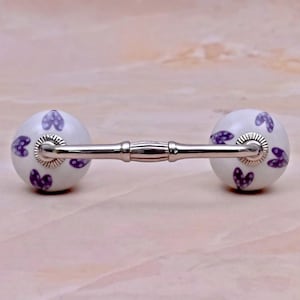Può includere: Una maniglia per mobili con due sfere in ceramica bianca decorate con cuori viola, collegate da una barra in metallo color argento lucido. Le estremità sferiche presentano una finitura liscia e lucida con piccoli cuori viola.
