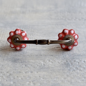 May include: A red and white polka dot ceramic drawer pull with a brass bar connecting the two knobs.