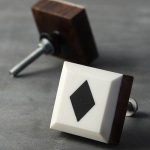 Könnte beinhalten: Zwei quadratische Schrankknöpfe mit Holzsockel und weißer Harzplatte. Ein Knopf hat eine schwarze Diamantform.