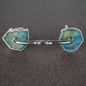 May include: A silver cabinet pull with two blue and green agate stone knobs. The knobs have a rough, natural edge.