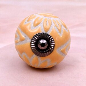 May include: A spherical ceramic cabinet knob featuring a raised, light orange floral pattern against a white background. The centre of the knob has a circular, metallic silver-toned hardware fitting with a ridged edge and a central screw hole.