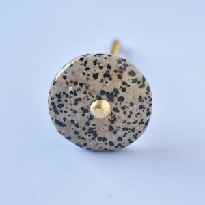 Op de afbeelding: Een ronde, gevlekte, grijze en zwarte knop met een gouden accent. De knop is gemaakt van steen en heeft een rustieke uitstraling.