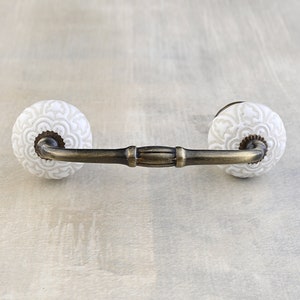 May include: A white ceramic drawer pull with a decorative floral design and a brass bar connecting the two knobs.