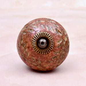 May include: A spherical cabinet knob with a textured, mottled surface in shades of salmon pink and beige. The center features a circular, fluted brass-toned metal plate surrounding a small, dark, recessed screw hole for mounting purposes.