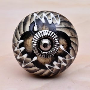 May include: A round ceramic cabinet knob featuring a swirling black and beige pattern with raised white zigzag lines. The centre of the knob is adorned with a circular, fluted silver-toned metal hardware piece with a small central screw head.