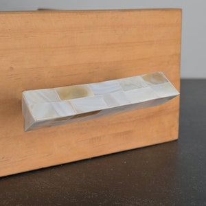 Könnte beinhalten: Ein gebogener Schubladenzug aus kleinen, quadratischen Fliesen in Weiß und Beige. Der Zug ist an einer hellbraunen Holzklappe montiert.