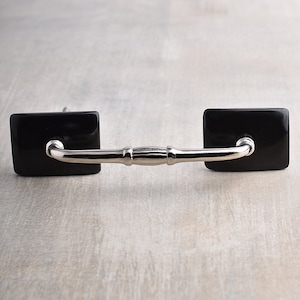 May include: Black cabinet hardware with a silver metal pull. The hardware has square black ceramic ends.