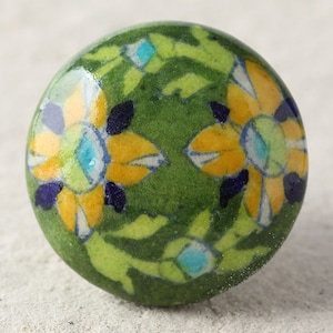 May include: A green ceramic knob with a floral design. The flowers are yellow with blue and white accents. The knob is round and has a smooth surface.