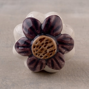 Puede incluir: Un pomo de cerámica blanca en forma de flor con un diseño de flor morada y un centro marrón. El pomo es perfecto para agregar un toque de encanto rústico a cualquier armario o cajón.