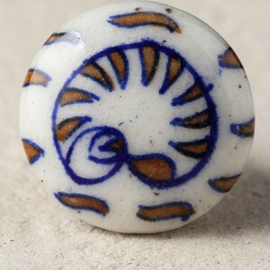 May include: A white ceramic knob with a blue and brown swirl design. The knob is round and has a smooth surface.