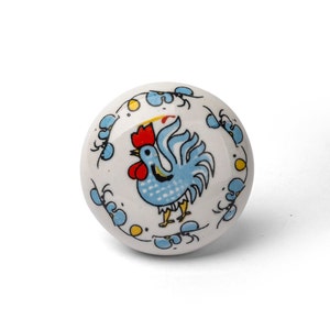 Puede incluir: Pomo de cerámica blanco con un diseño de gallo azul. El gallo tiene una cresta y barbillas rojas, y está rodeado de acentos florales azules y amarillos.