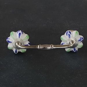 Könnte beinhalten: Ein weißer Keramik-Schubladengriff mit blauen und grünen Blumenmustern. Der Griff hat eine silberne Metallstange, die die beiden Blumenknöpfe verbindet.