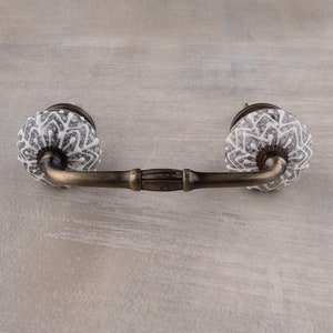 May include: A white ceramic drawer pull with a decorative pattern and a brass bar connecting the two knobs.
