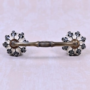 May include: A drawer pull featuring a horizontal antique brass metal handle connected to two round, fluted ceramic knobs. The knobs are white with a black botanical leaf pattern and are accented with brass-toned circular metal bases.