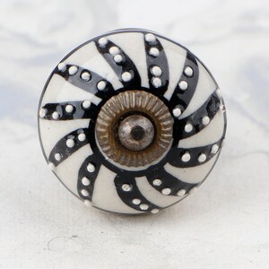 May include: A white ceramic knob with a black and white swirl design and black dots. The knob has a brass centre.