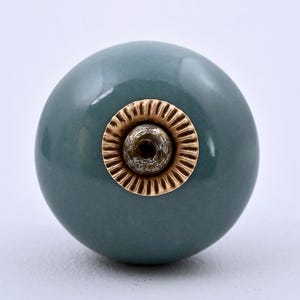 Puede incluir: Un pomo de mueble redondo de cerámica en color verde salvia liso, con una pieza central de metal en tono dorado. El componente metálico presenta un borde circular estriado que rodea un pequeño punto central oscuro y texturizado, creando contraste.