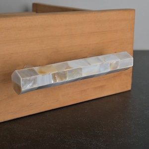 May include: A white and beige shell mosaic drawer pull with a rectangular shape. The pull is mounted on a wooden drawer.
