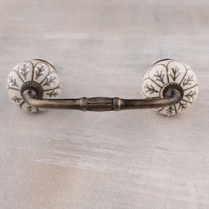 Puede incluir: Un pomo de armario de cerámica blanca con un diseño floral verde y un brazo de metal bronce. El pomo está unido al brazo con un tornillo.