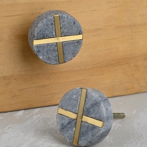 Może przedstawiać: Dwa okrągłe, szare marmurowe pokrętła do szafki z wzorem złotego krzyża. Pokrętła mają średnicę około 3,8 cm.
