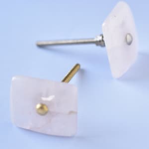 Könnte beinhalten: Zwei quadratische rosa Schrankknöpfe mit goldenen Akzenten und silbernen Schrauben.