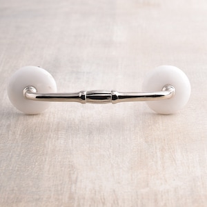 Op de afbeelding: Witte keramische ladegreep met een zilveren metalen balk die de twee knoppen verbindt.