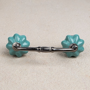 May include: A cabinet pull featuring a dark metal handle and turquoise ceramic flower-shaped knobs. The knobs have a crackle glaze finish. Suitable for drawers and cabinets.