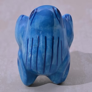 Könnte beinhalten: Eine blaue Keramik-Froschfigur mit blauen und weißen Streifen. Der Frosch sitzt auf einer weißen Oberfläche.
