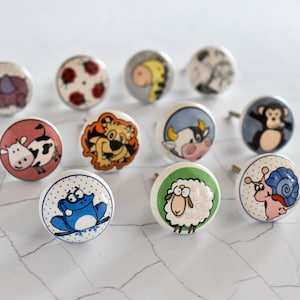 Op de afbeelding: Een set van 10 kleurrijke keramische knoppen met verschillende dierenontwerpen, waaronder een koe, een kikker, een slak, een schaap, een aap, een lieveheersbeestje, een tijger en een koe. De knoppen zijn perfect om meubels of kasten een speelse touch te geven.